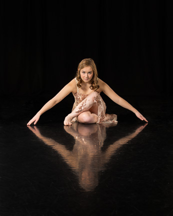 Dancer sitting crossed-legged on reflective black floor with arms outstretched, fingertips touching floor creating a unique sculptured look.